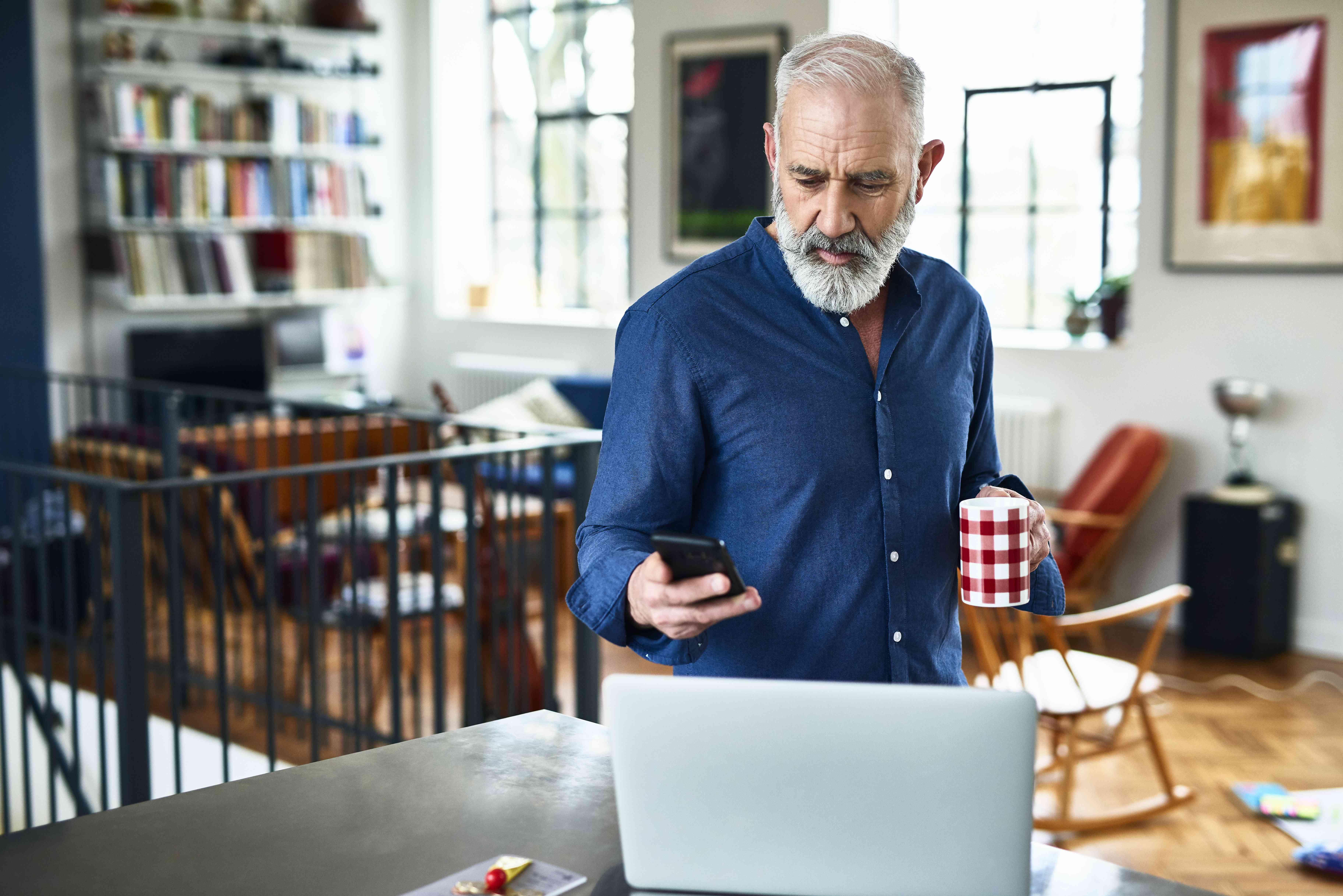 man looks at phone and laptop while holding coffee cup