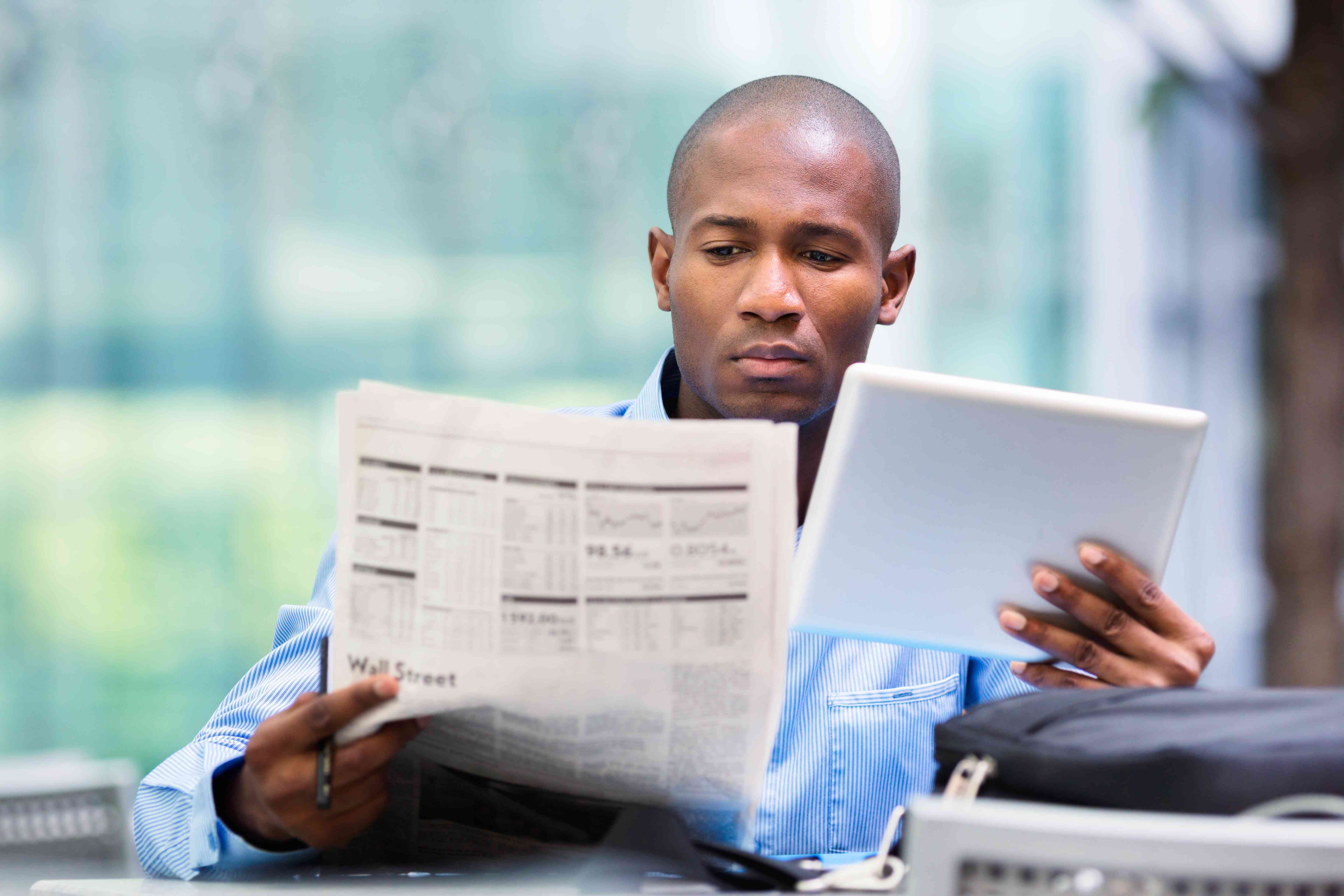 Investor reading over newspaper while holding a tablet