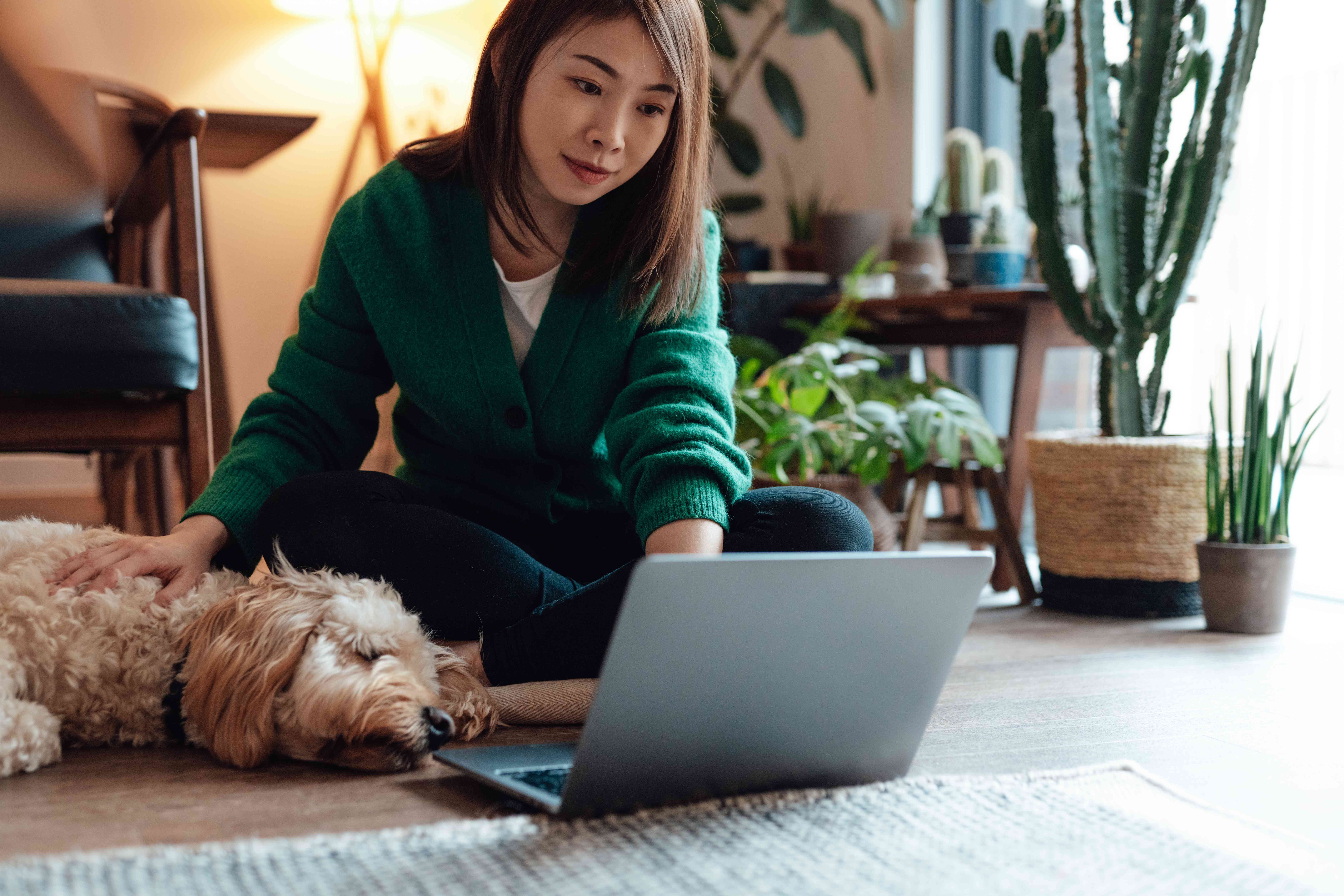 Woman patting a dog while using a laptop computer and seated on the floor.
