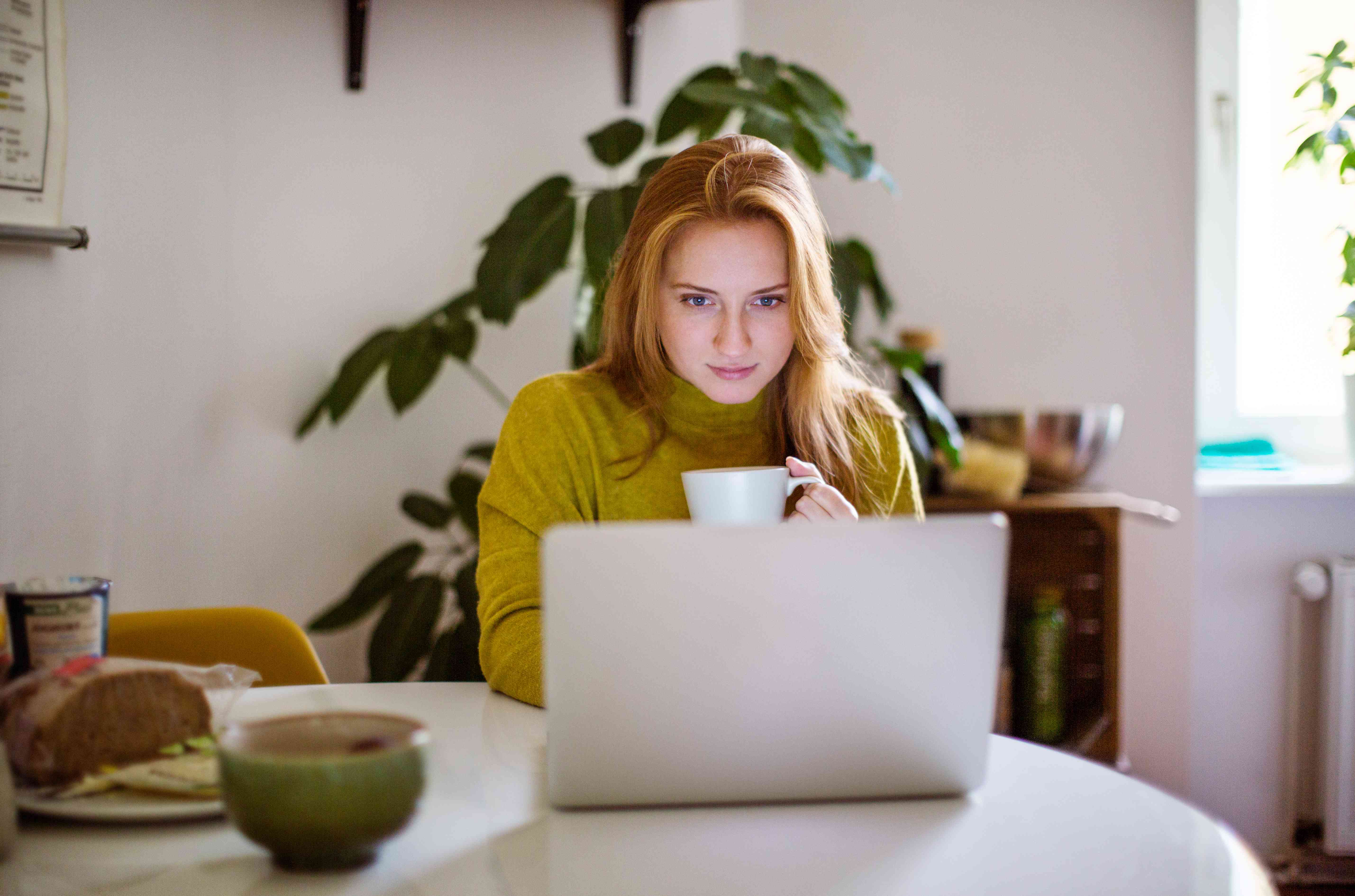Young woman sitting at table looking at her laptop and drinking coffee.